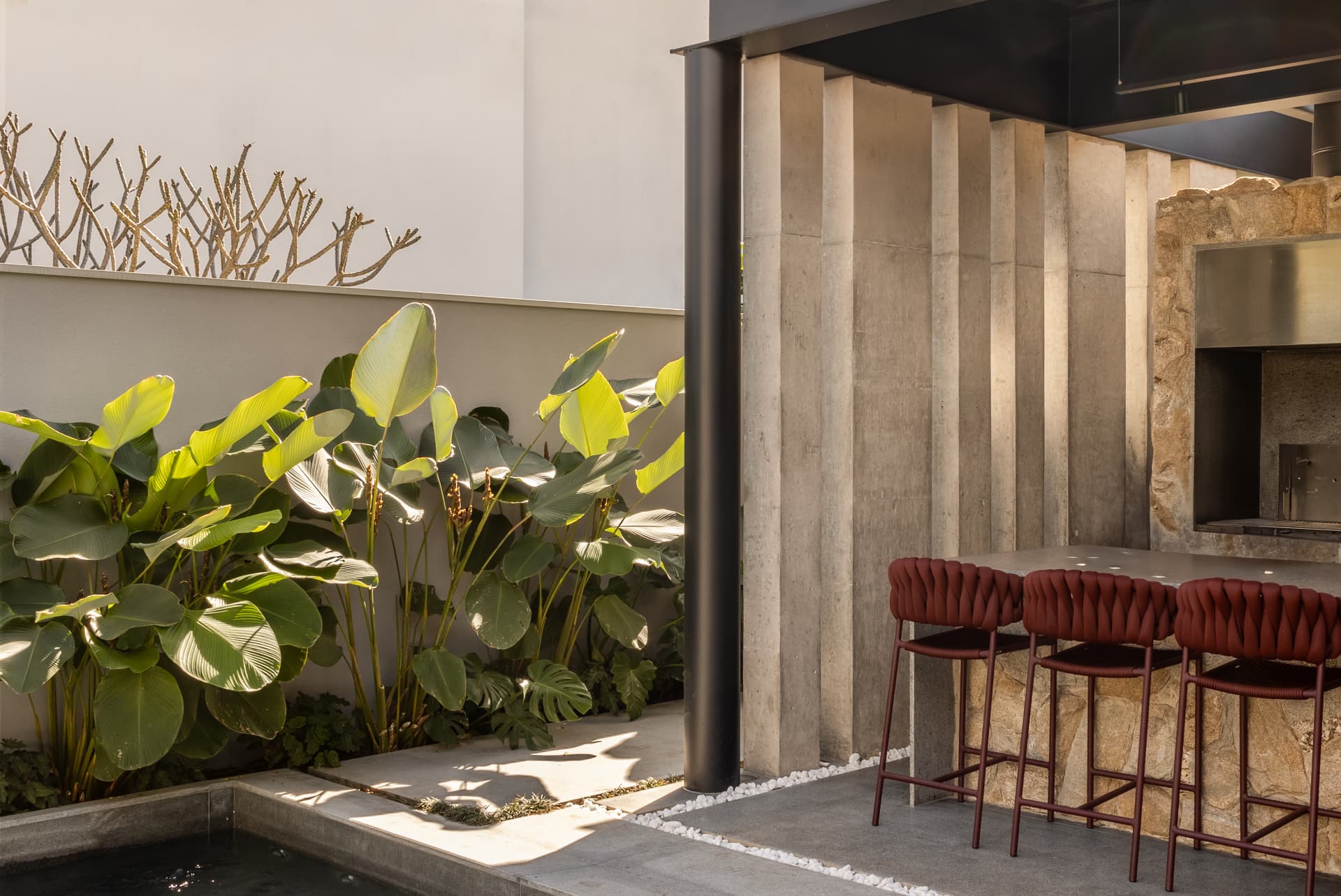 Área gourmet com churrasqueira de pedra, banquetas e folhagens ao sol