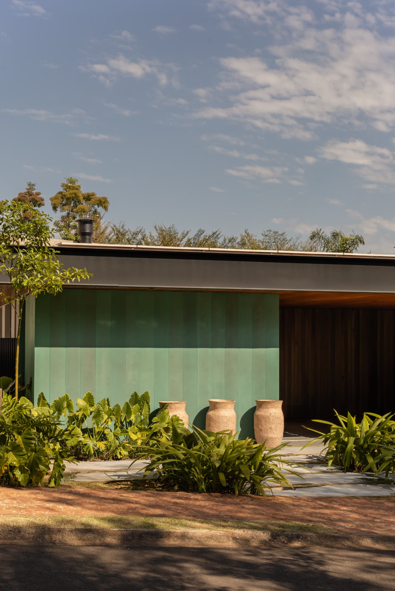 Fachada com painel verde, três vasos de pedra e maciço de folhagens sob céu azul